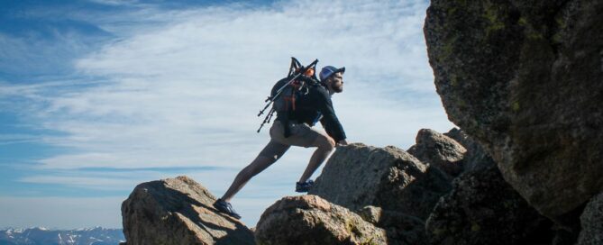 Man climbing rocks outdoors, with blue skies above him and a backpack on his back.