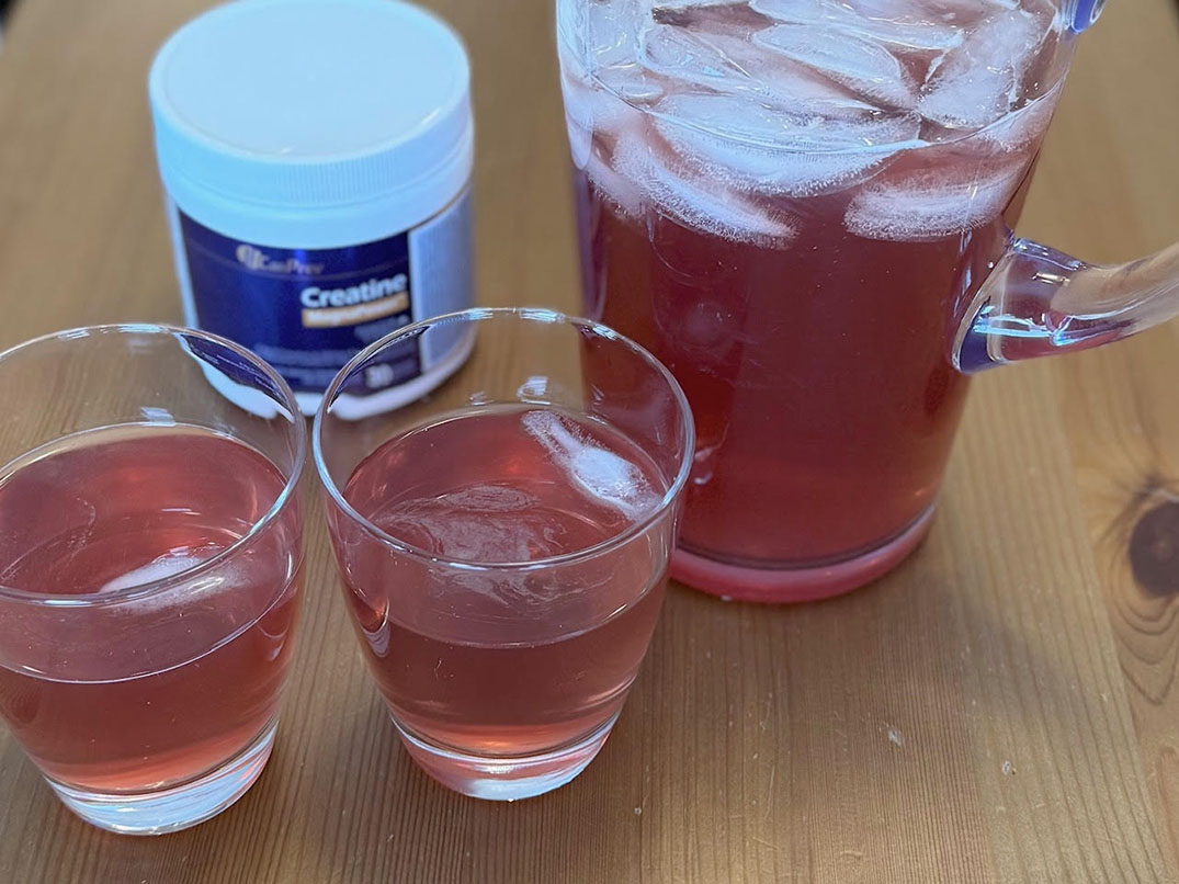 Two small glasses and a pitcher with a pink drink on a wooden table; a tub labeled Creatine sits in the background.