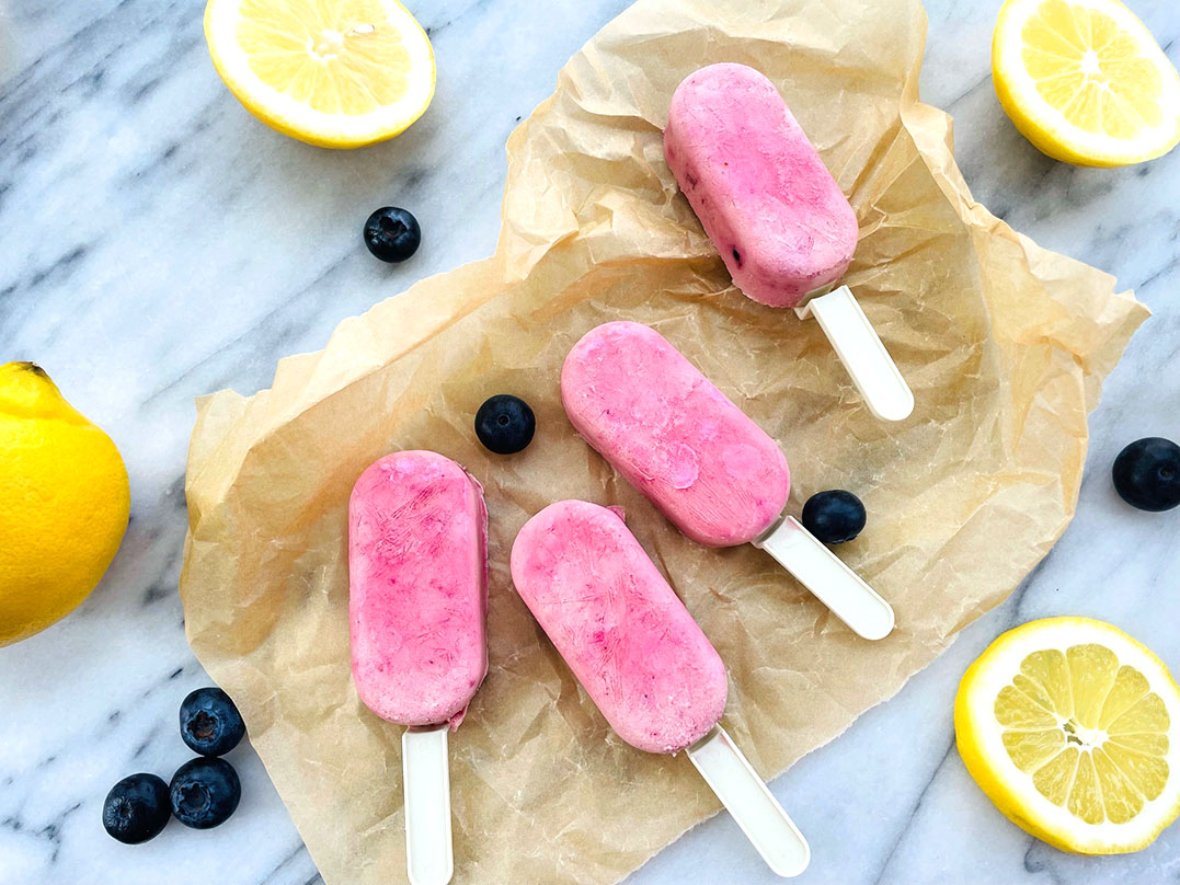 Four pink popsicles on white sticks rest on crumpled parchment, surrounded by blueberries and lemon wedges on a marble surface (top-down shot).